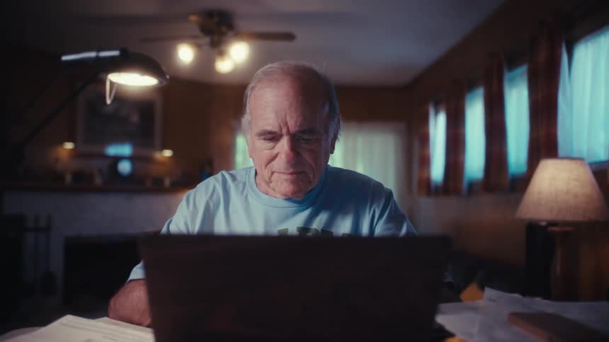 An elderly army veteran finishes with paperwork, opens up his laptop, and begins working at his desk in a warmly lit room