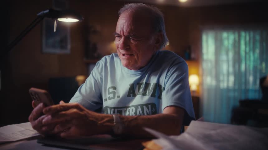 An elderly U.S. Army veteran smiles as he looks at his smartphone while sitting at his desk at home