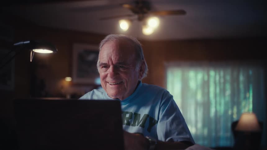 An elderly US Army veteran joyfully opens his laptop at his desk in his dimly lit living room