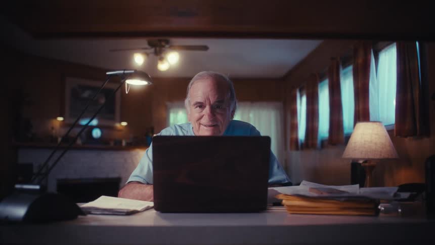 An elderly Army veteran's face lights up with a warm smile as he opens his laptop at his desk to connect with someone online