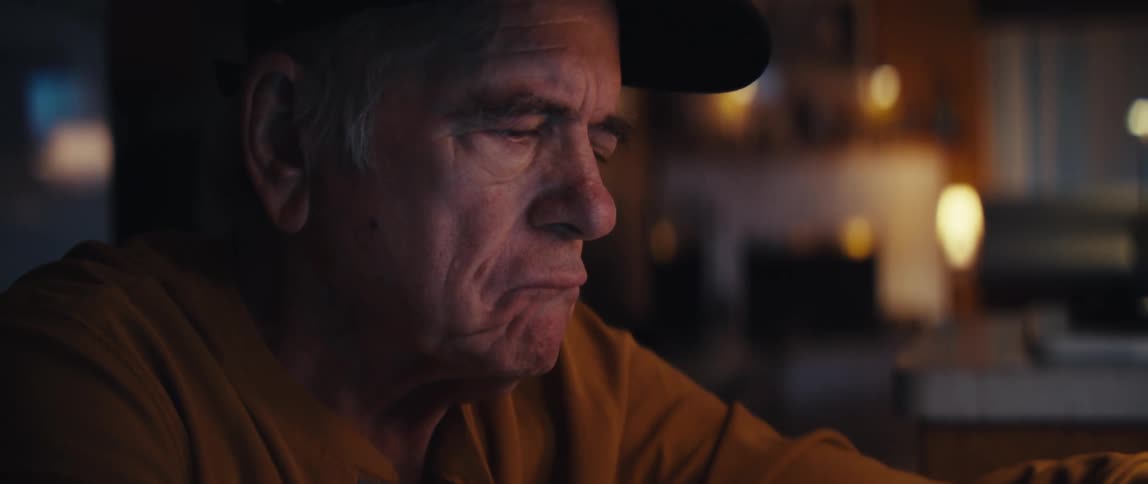 A focused elderly man wearing a hat carefully calculates his bills late at night in a dimly lit room