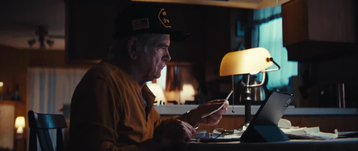 An elderly US Army veteran with a serious expression sits at his kitchen table at night writing in a notebook