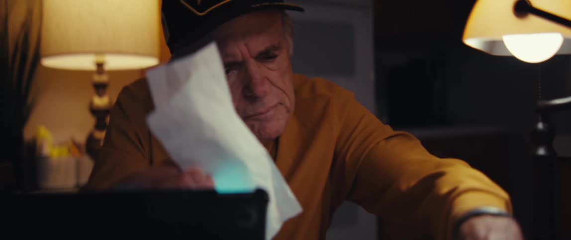 An elderly US Army veteran with a worried expression sorts through bills and receipts at his desk at night