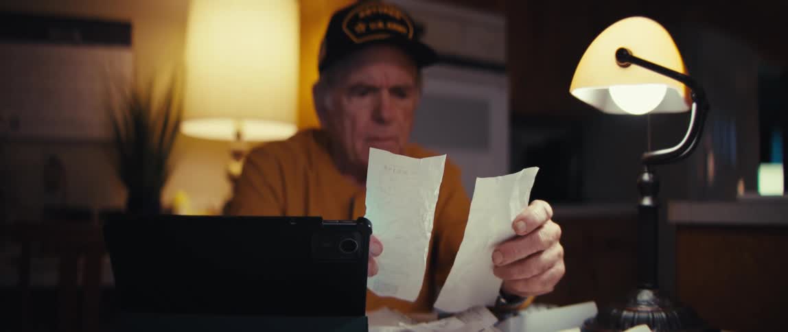 An elderly retired army veteran sits at a table late at night, looking over receipts with a worried expression as he manages his finances