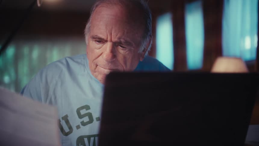 Slow zoom in on an elderly man looking over papers at his desk at night, his serious expression turning into a heartwarming smile