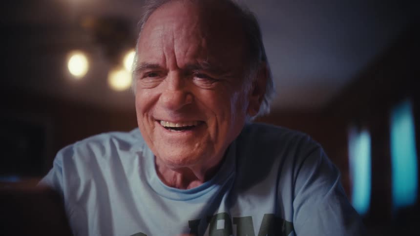 An elderly army veteran smiles warmly as he scrolls through his smartphone at his desk late at night