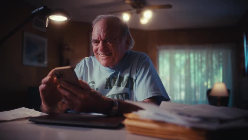 A joyful elderly Army veteran sits at his desk at home and laughs heartily while looking at his smartphone