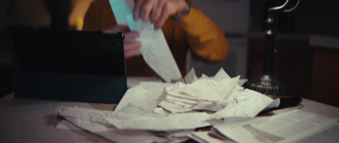 An elderly Army veteran with a worried expression sorts through a messy pile of receipts and bills at his kitchen table late at night