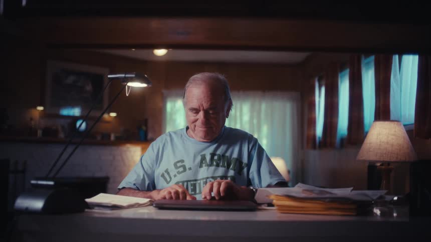 An elderly U.S. Army veteran smiles warmly as he opens his laptop at his desk in a cozy, dimly lit home office