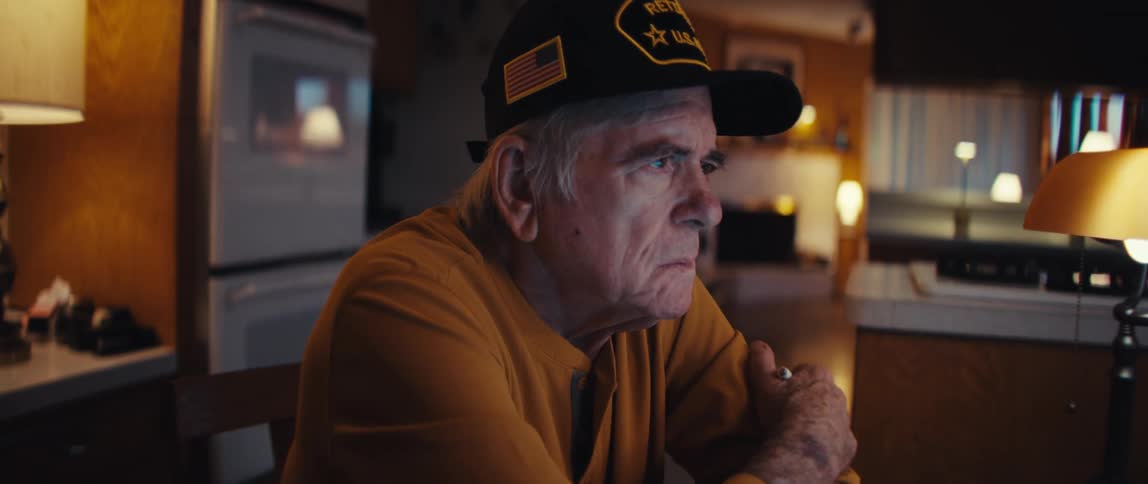An elderly retired U.S. Army veteran with a concerned expression sits at his kitchen table contemplating his finances at night