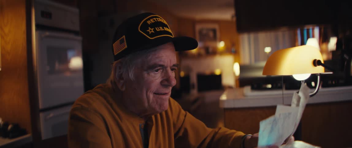 An elderly retired U.S. Army veteran smiles warmly as he looks over papers at his kitchen table in the evening