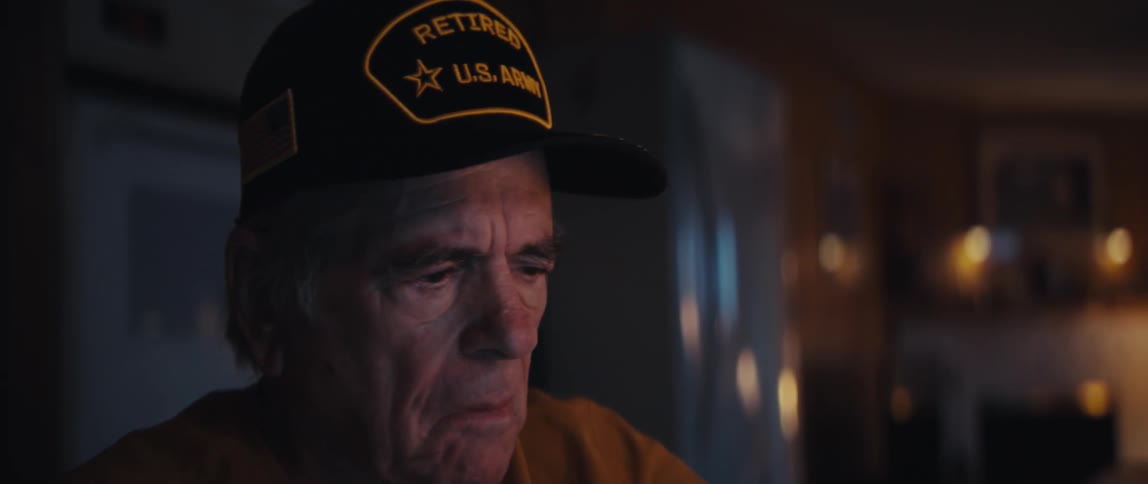 A close-up shot of a thoughtful elderly man, a retired U.S. Army veteran, looking down with a sad expression in a dimly lit room
