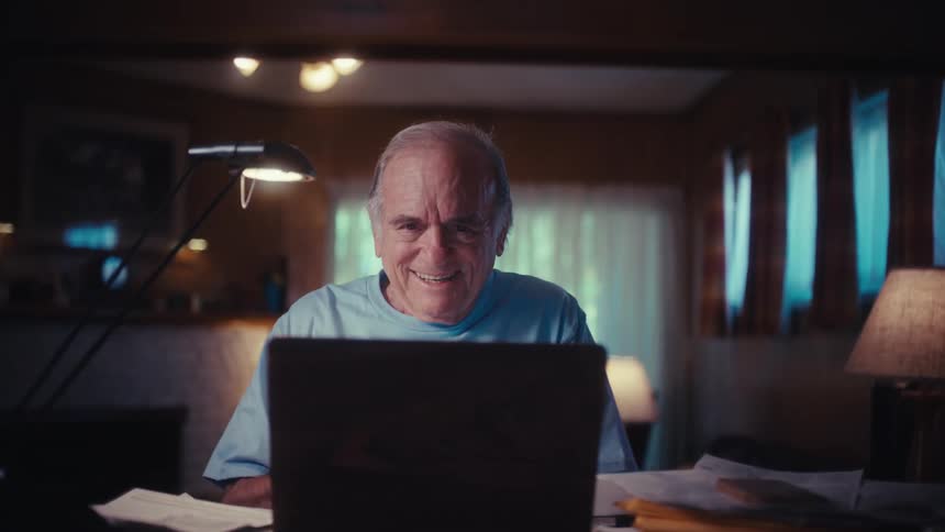 A smiling elderly US Army veteran opens his laptop at his desk in a dimly lit room, his face filled with joy and surprise as he looks at the screen