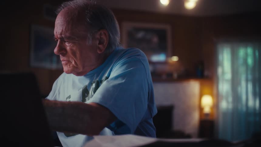 A concerned elderly U.S. Army veteran reviews documents and uses his laptop at his desk in a dimly lit room