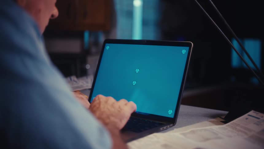 An over-the-shoulder view of an elderly man typing on a laptop with a green screen, surrounded by paperwork at his desk at night