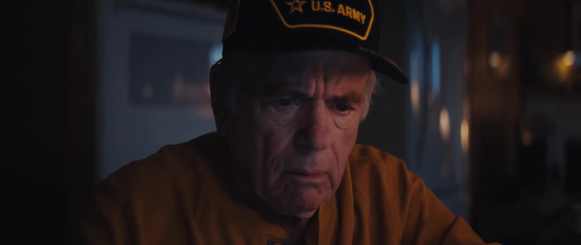 A pensive elderly veteran wearing a U.S. Army hat stares intently at a screen in a dimly lit room at night