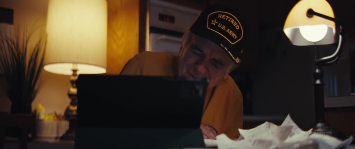 An elderly retired U.S. Army veteran smiles and laughs while video chatting with loved ones on his tablet in a warmly lit home