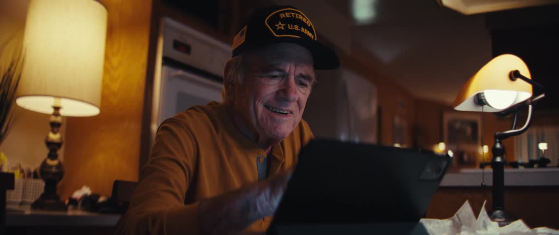 An elderly U.S. Army veteran smiles warmly while using a tablet computer in his cozy home at night