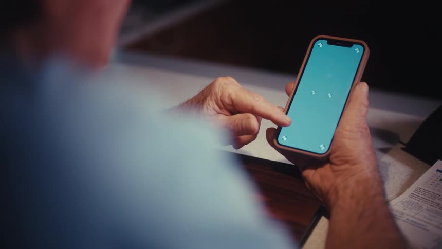 An over-the-shoulder view shows an elderly man's hands as he scrolls on a smartphone with a green screen
