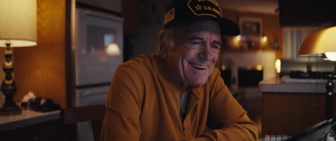 A happy elderly veteran wearing a U.S. Army hat smiles warmly while using a tablet at his kitchen table at night