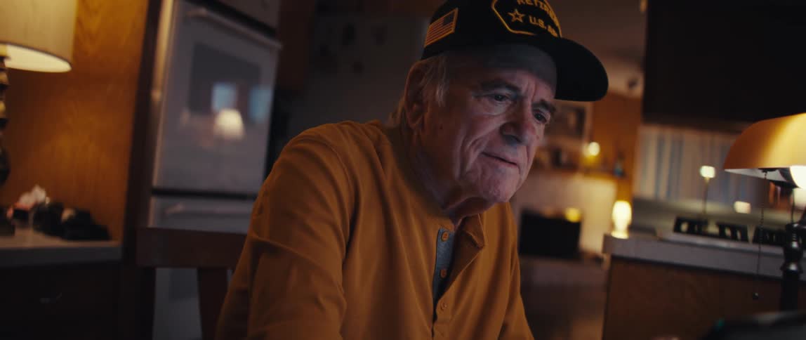 A retired US Army veteran sits at his kitchen table at night, a warm smile spreading across his face as he looks at his tablet
