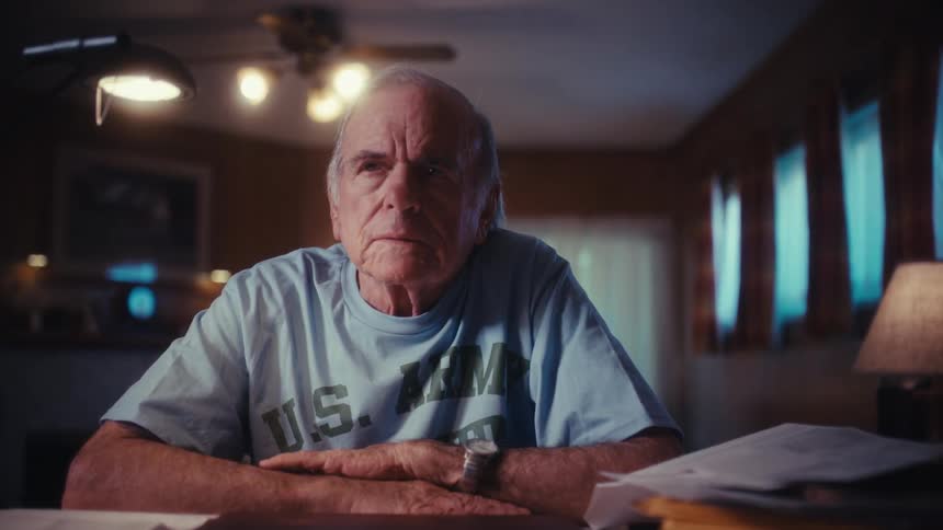 A thoughtful elderly army veteran closes his laptop in frustration and sits at his desk at night, lost in thought