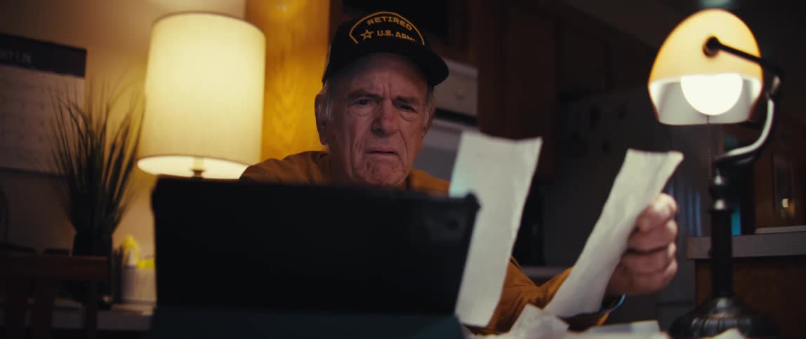 A worried elderly veteran sits at a table covered in receipts, anxiously reviewing his finances late at night