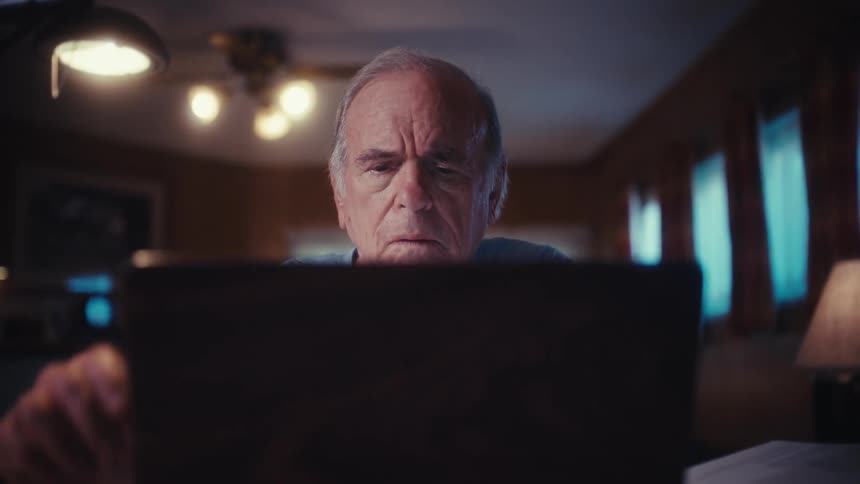 A worried elderly US Army veteran closes his laptop after working late at night in his dimly lit home office