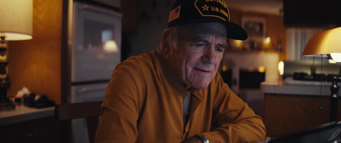 An elderly retired U.S. Army veteran sits at his kitchen table at night, smiling fondly while looking at a tablet