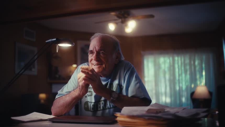 An elderly veteran closes his laptop and smiles as he fondly reminisces while sitting at his desk in a dimly lit room