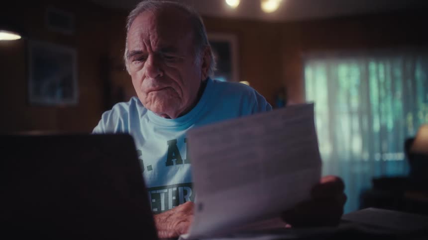 A concerned elderly Army veteran reviews his bills and documents on a laptop at his desk in a dimly lit room