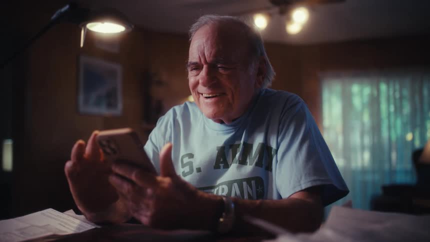 A joyful elderly U.S. Army veteran laughs while looking at his smartphone at a desk in his home