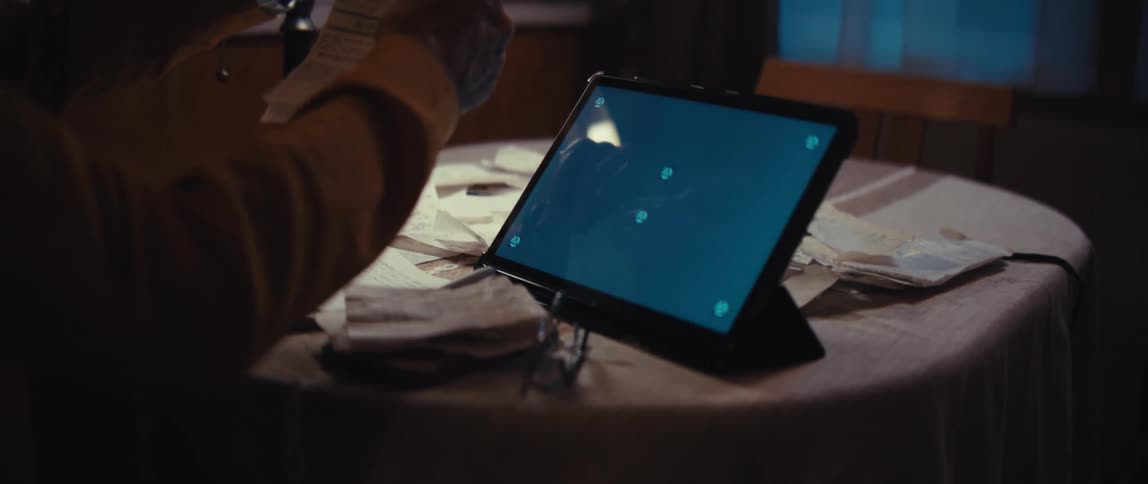 A person meticulously reviews receipts at a dimly lit table, using a calculator and a tablet with a green screen for financial management