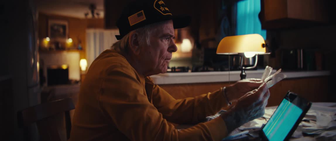 A worried elderly US Army veteran sits at his kitchen table late at night, trying to manage his finances and pay bills using a tablet