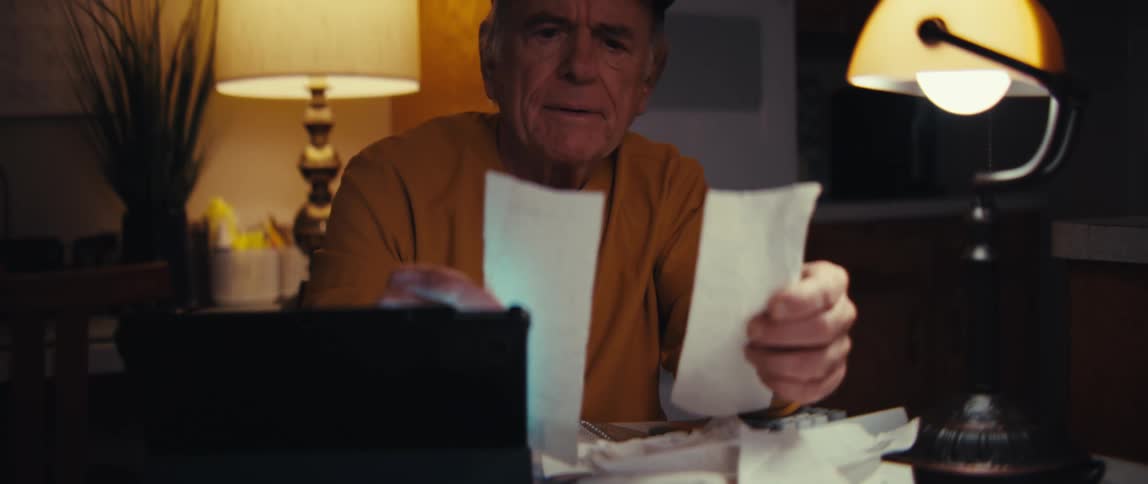 An elderly retired U.S. Army veteran sits at his kitchen table late at night, overwhelmed while sorting through a mountain of receipts