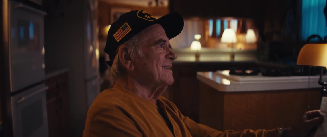 An elderly veteran wearing a hat with an American flag smiles warmly while watching something off-camera in his cozy home