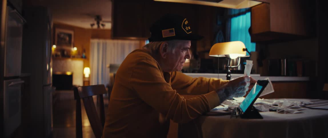 Smiling with contentment, an elderly veteran uses a tablet to manage his finances at his kitchen table in the evening