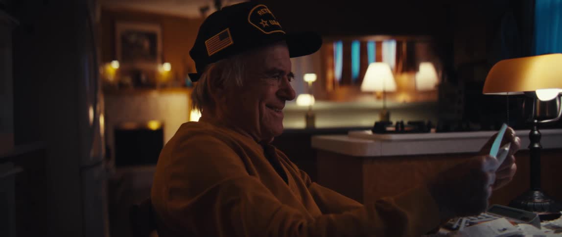A retired U.S. Army veteran sits at his kitchen table at night, smiling and laughing as he reads a letter that brings him joy