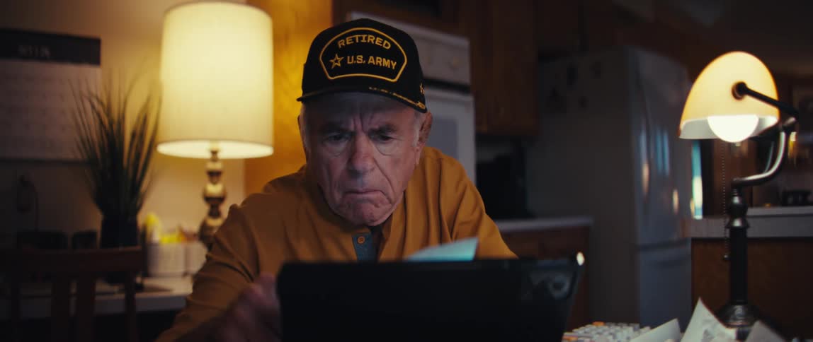 A concerned elderly army veteran sits at his dimly lit kitchen table, reviewing his finances with a tablet and a pile of receipts