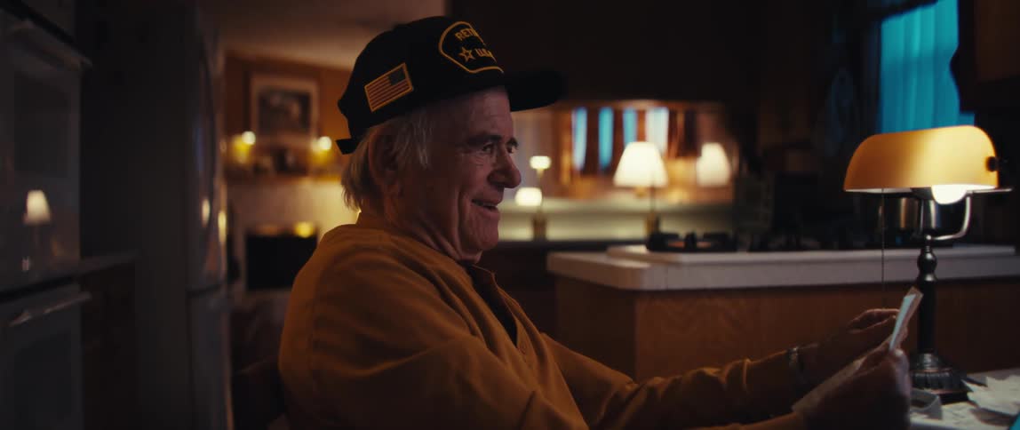 A retired US Army veteran sits at his kitchen table at night, smiling fondly as he reminisces over an old photograph