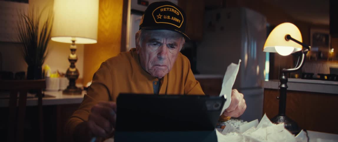 An elderly retired Army veteran with a concerned look manages his finances at night, using a tablet and looking over receipts