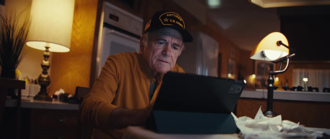 An elderly U.S. Army veteran sits at a table in his home at night, smiling as he looks at his tablet