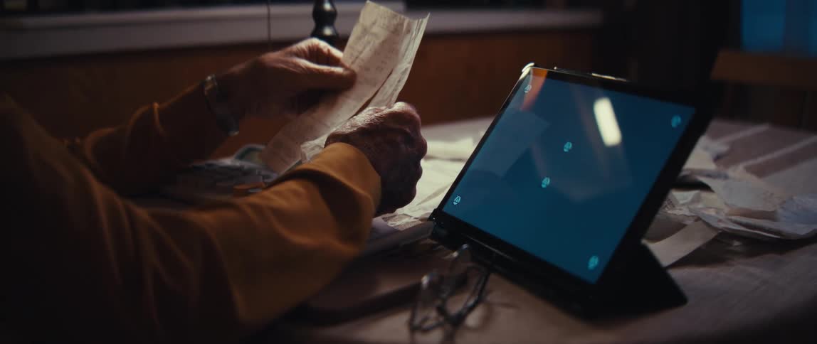An elderly person carefully reviews receipts at a cluttered desk next to a tablet with a green screen