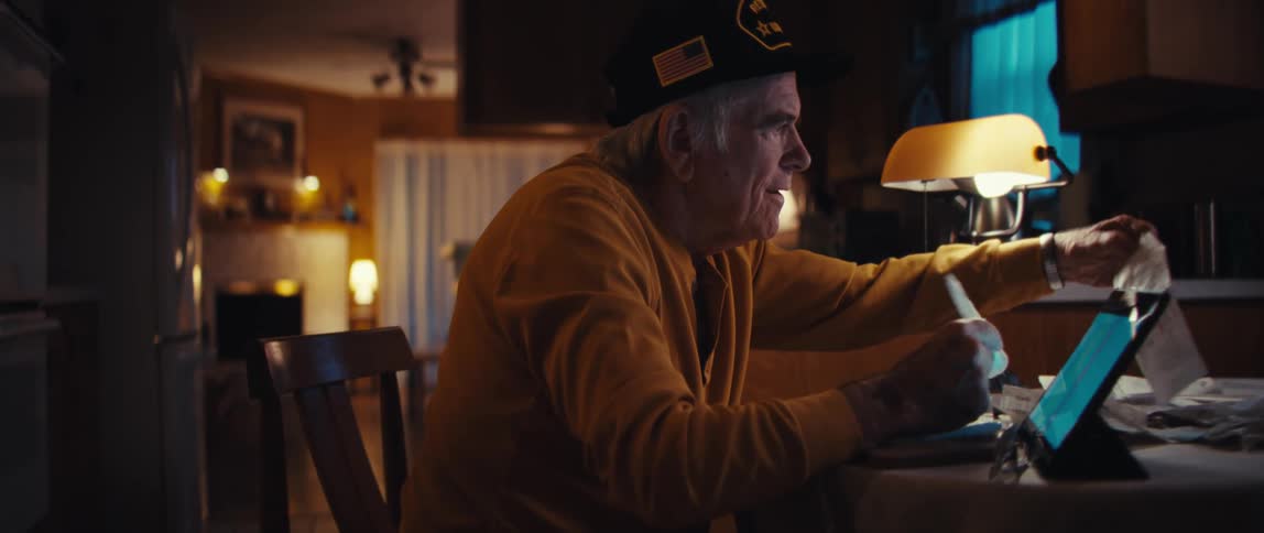 A retired U.S. Army veteran smiles warmly while reviewing receipts and using a tablet at his kitchen table late at night