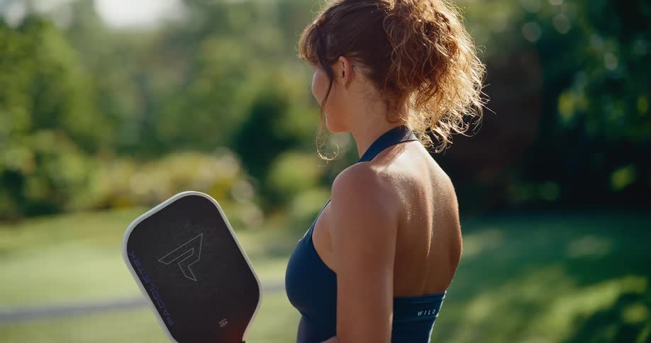 A smiling woman in athletic wear holds a pickleball paddle on a sunny outdoor court, turning her back to the camera in slow motion