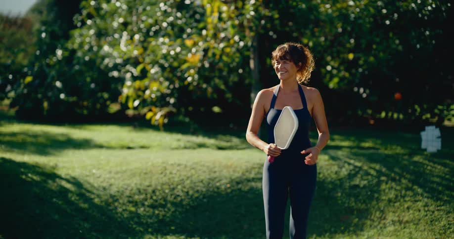 A smiling woman in sportswear strolls across a lush green lawn on a sunny day, holding a pickleball paddle