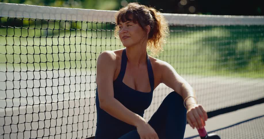 A beautiful female athlete relaxes against the net on a pickleball court, enjoying the warm sunshine