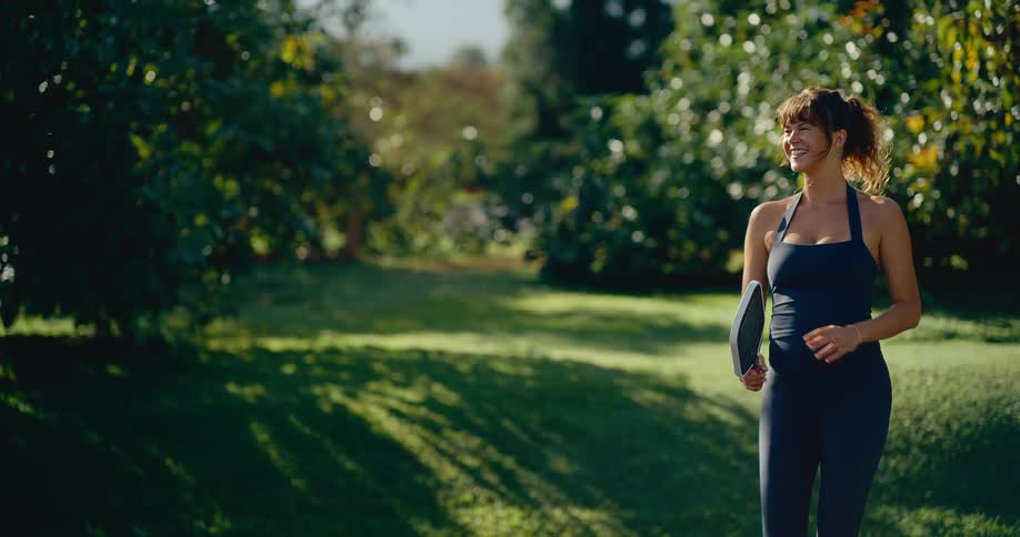A smiling woman in athletic wear walks through a sunny park before hitting a pickleball in slow motion