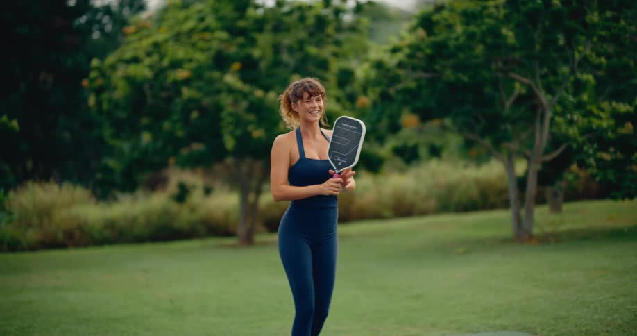 An athletic woman joyfully plays pickleball, serving the ball in slow motion in a beautiful outdoor setting