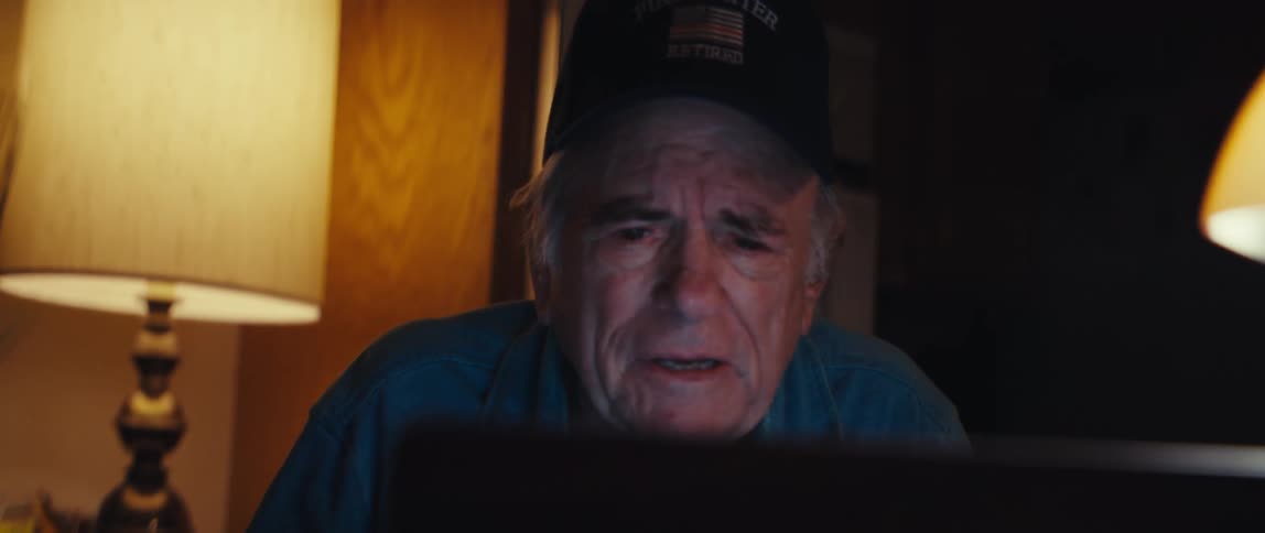 Close-up of a teary-eyed elderly man in a retired firefighter hat looking at a laptop screen in a dark room